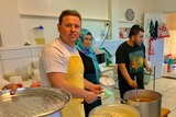One man looking at camera while dishing up rice in the mosque's kitchen, and a man and a woman serving food behind him