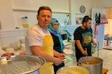 One man looking at camera while dishing up rice in the mosque's kitchen, and a man and a woman serving food behind him