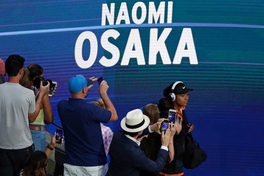 A tennis player walks onto court wearing headphones as fans take photos of her.