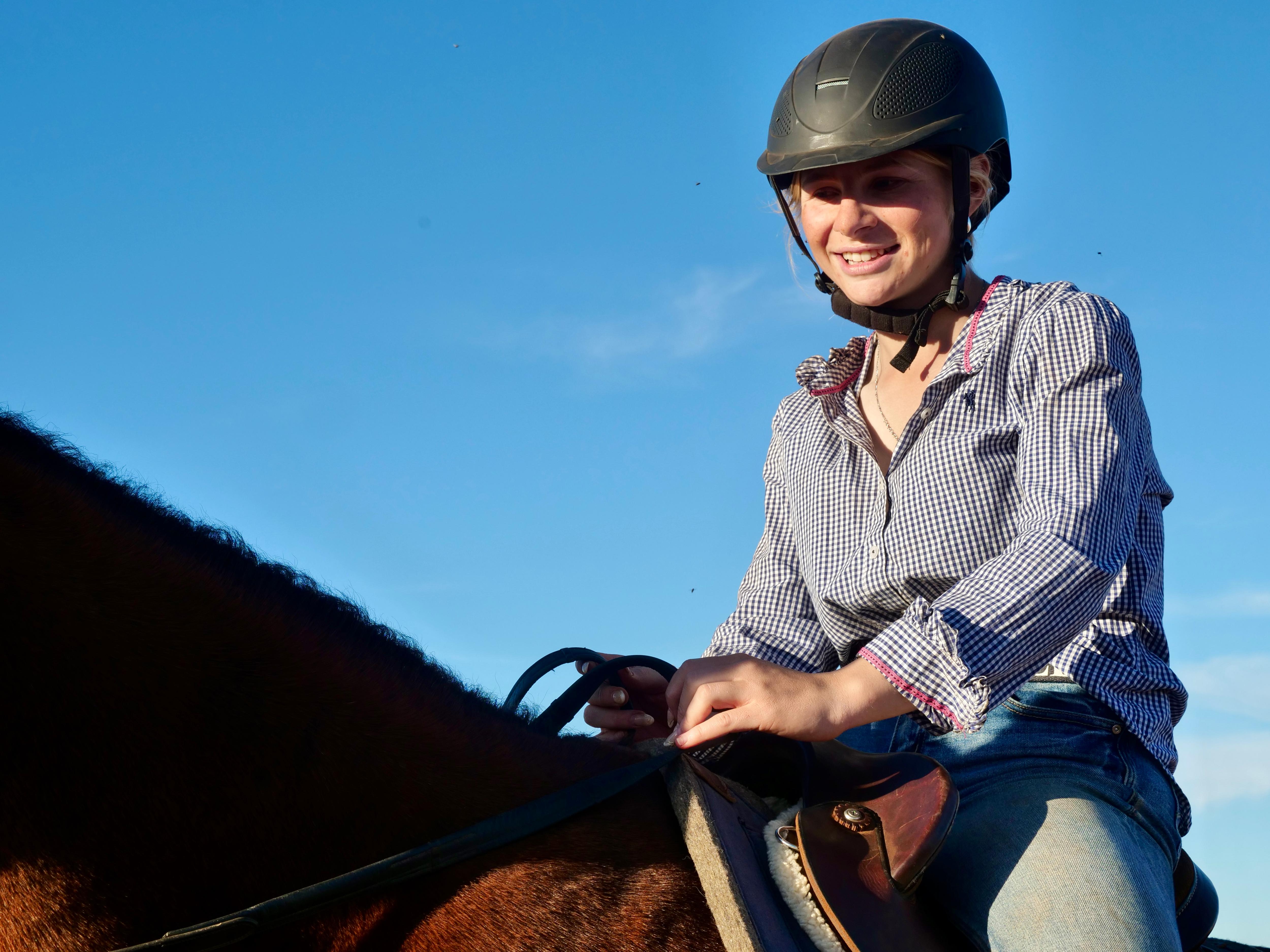 a large brown horse with a girl in a helmet and a blue shirt and jeans sitting in a saddle on the back of the horse