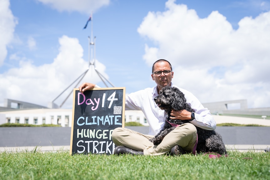 A man sits outside Parliament House with a dog and a sign that says 'Day 14 Climate Hunger Strike'.