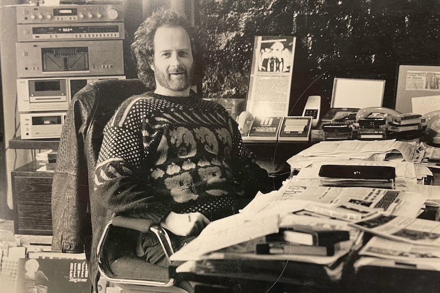 A black and white photo of Michael Gudinski sat at a cluttered office desk wearing a knitted jumper and jeans