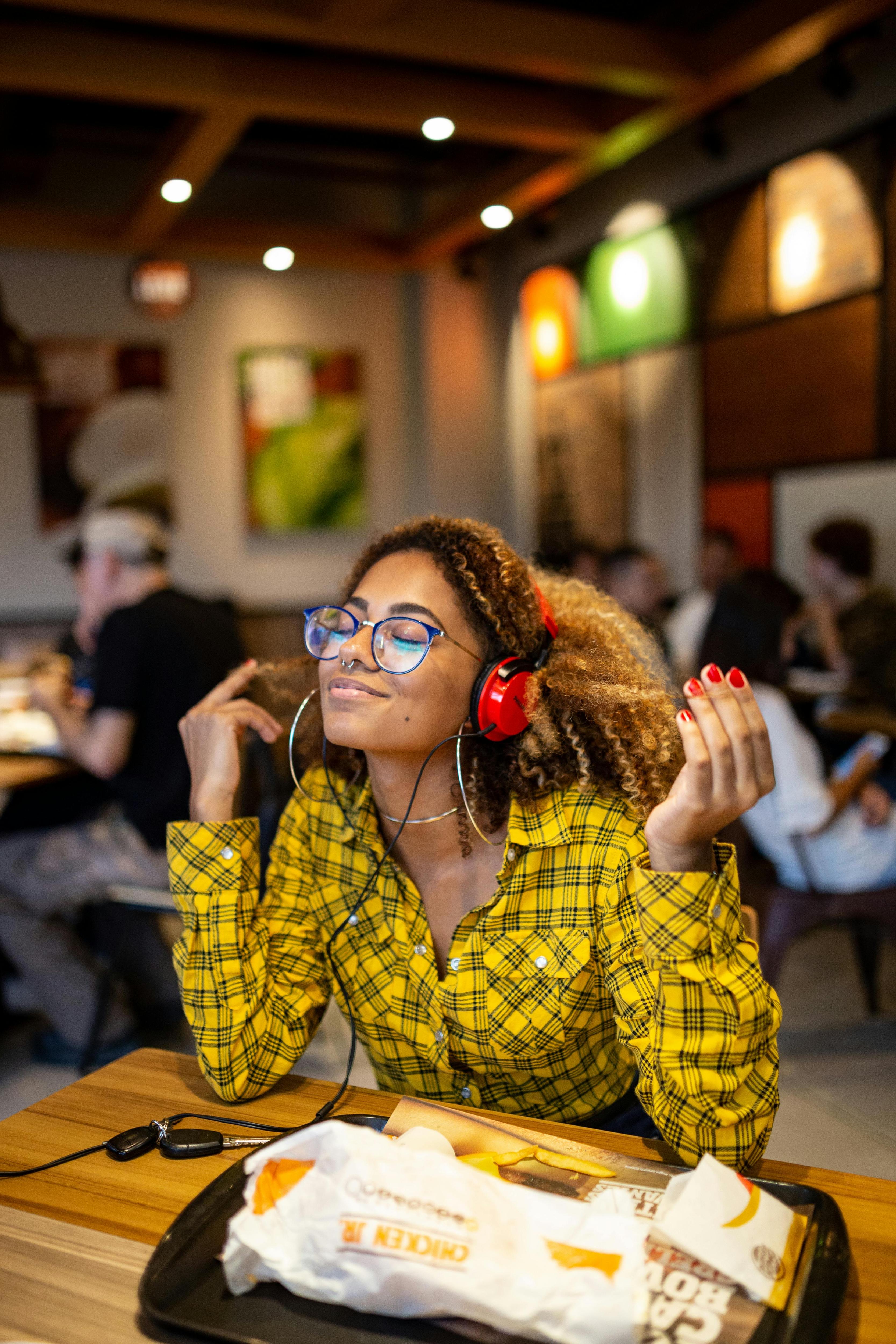 A woman with curly hair and a yellow and black check top listens to music through red headphones in a restaurant
