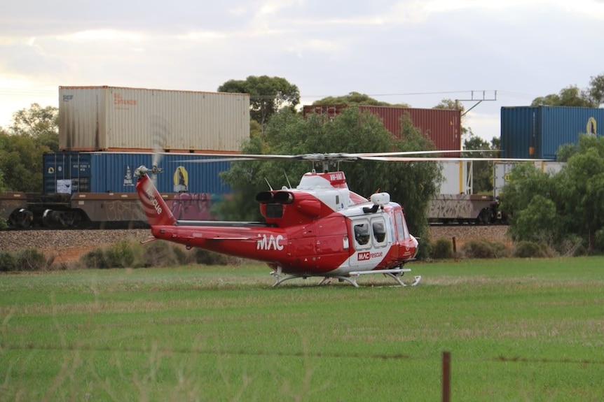 A red helicopter in a field in front of a freight train.