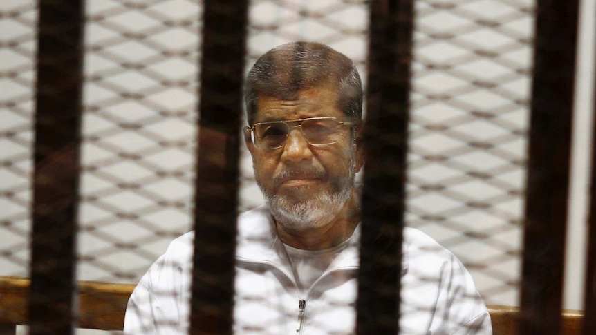 Mohammed Morsi wears a white prison jumpsuit and sits behind a cage door