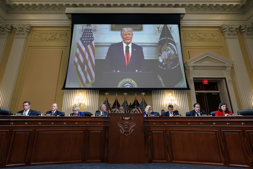 A row of people sitting in a room with a picture of Donald Trump behind them 
