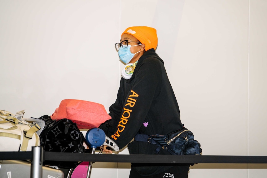 Naomi Osaka wheels her luggage wearing a black hoody and orange hat