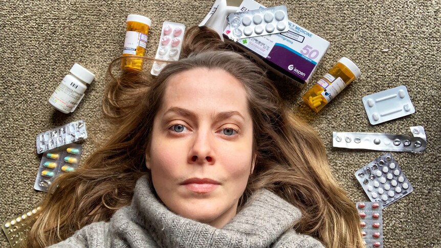 A woman with long brown hair in a grey turtleneck lays on the ground, surrounding by packets and bottles of pills
