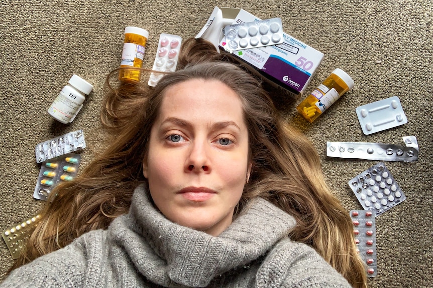 A woman with long brown hair in a grey turtleneck lays on the ground, surrounding by packets and bottles of pills