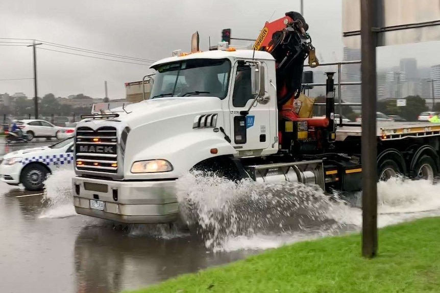 A freight truck drives through a flooded street spraying water out the sides of its tyres on a grey day.