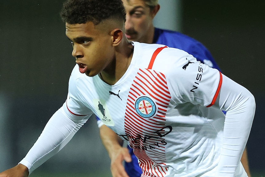 Raphael Borges Rodrigues runs, slightly hunched over looking forward with the ball at his feet and a defender behind him.