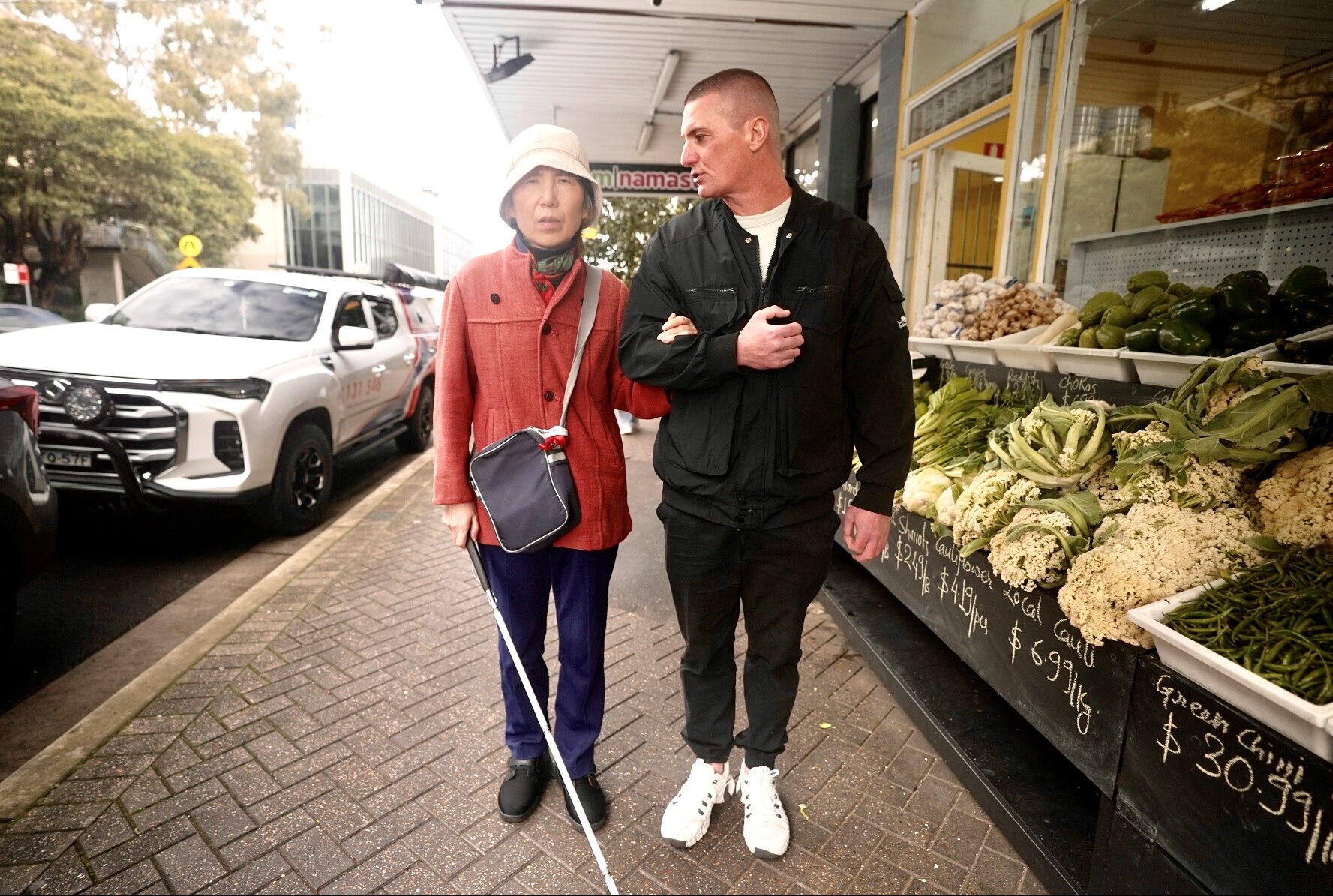 An elderly Asian woman, holding a white cane, and standing next to a larger, middle-aged Caucasian male.