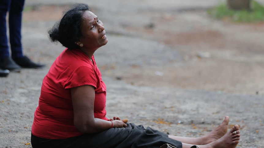 A woman in a red shirt sits on the ground looking off into the distance and sobbing.