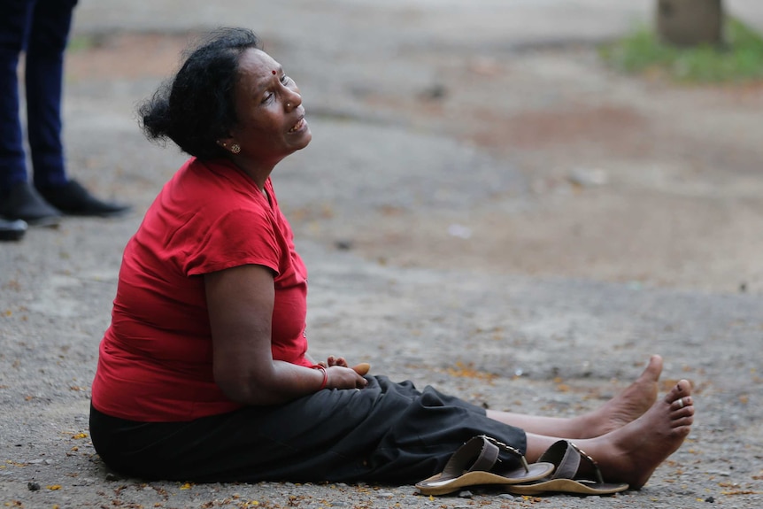 A woman in a red shirt sits on the ground looking off into the distance and sobbing.