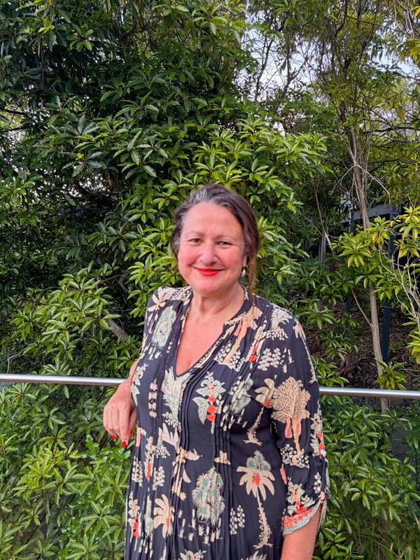 Una mujer sonriente de cabello oscuro con un top floral se apoya en la barandilla de un balcón con vista a un jardín verde.