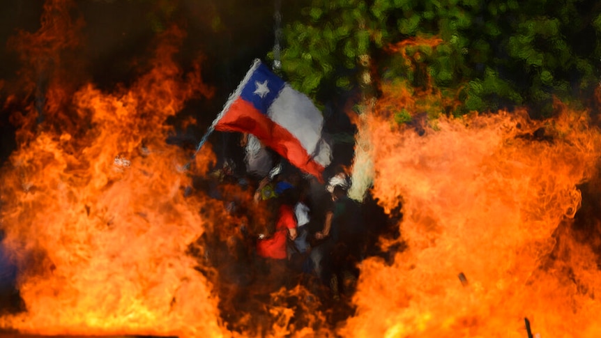 Viewing through a burning barricade fire, you view a Chilean flag being waved in the distance.