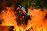 Viewing through a burning barricade fire, you view a Chilean flag being waved in the distance.