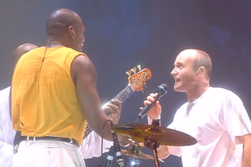 Phil Collins sings on stage near a drummer and bass player.