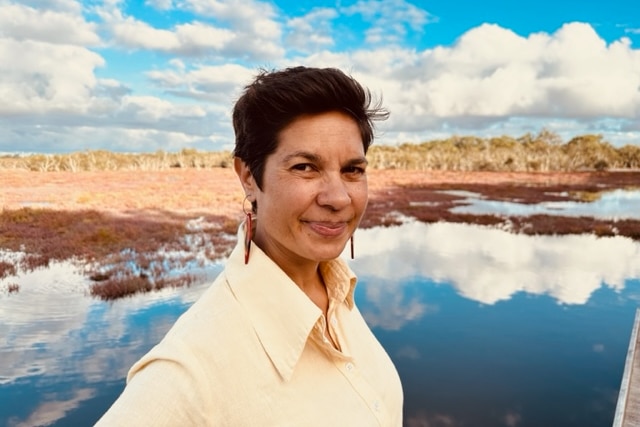 Narelda Jacobs, wearing a yellow shirt and short dark hair, poses in front of a river reflecting blue, cloudy skies