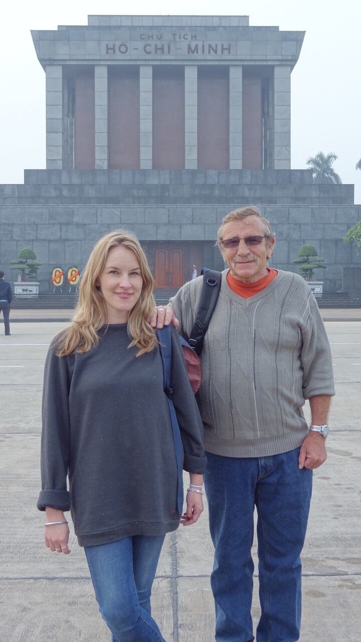 Man with arm on woman's shoulder standing in front of grand building with words, Ho Chi Minh.