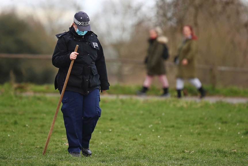 Un policier portant un masque facial et tenant un long bâton marche sur une zone herbeuse