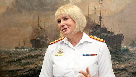 Woman in white uniform stands in front of painting of ship