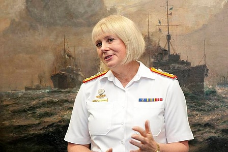 Woman in white uniform stands in front of painting of ship