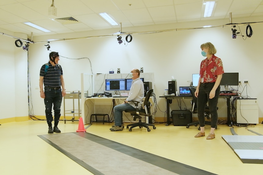 An elderly man wears a helmet fitted with sensors. He is facing two masked researchers. 