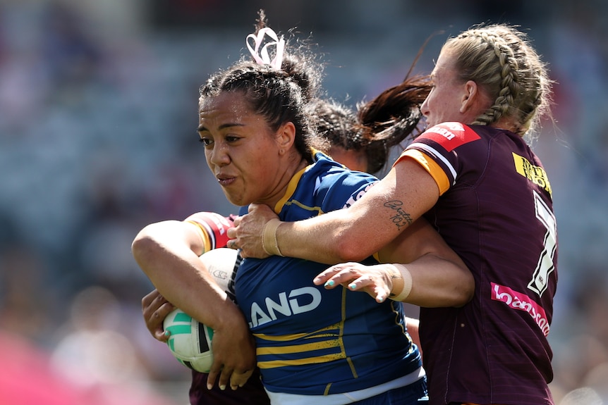 A Parramatta Eels NRLW player carries the ball as she is tackled by two Brisbane Broncos' opponents.