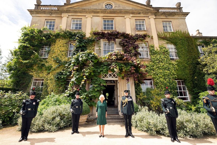 Camilla stands with a line of men in uniform outside an ornate house covered in vines and plants.