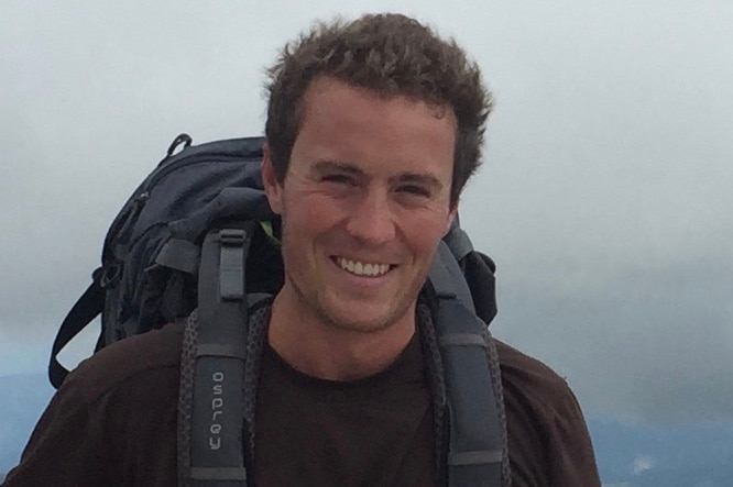 A young man wearing a hiking backpack smiles on a mountain summit, with clouds in the background.
