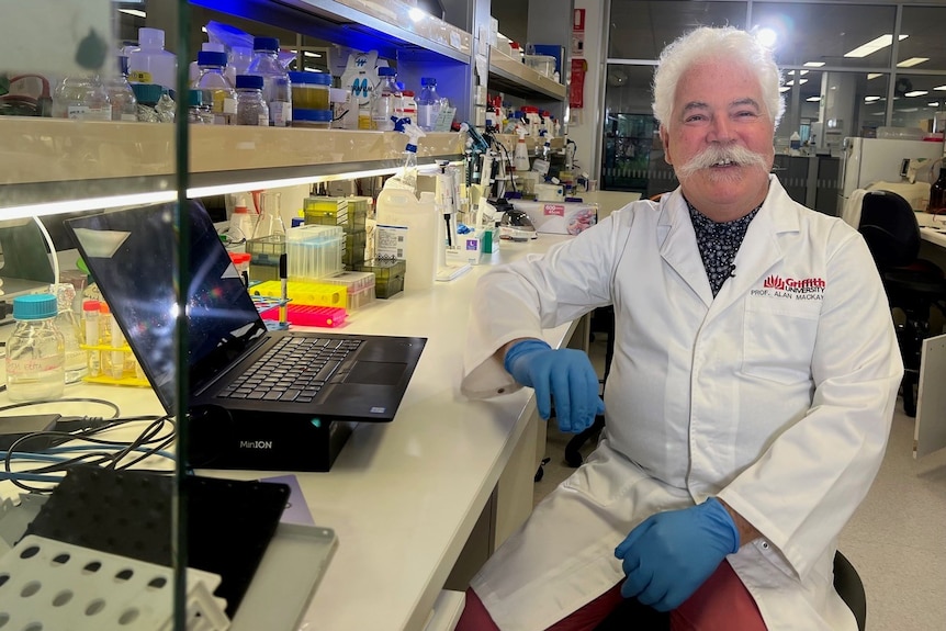 A man in a lab coat and gloves sits in a lab by his desk, smiling.