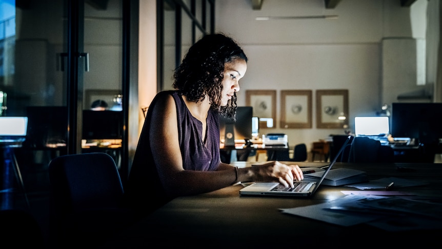 Woman with dark curly shoulder length hair and light brown skin sits in front of a laptop in an office late at night 