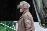 A woman with grey hair wears a surgical mask while walking past a tram