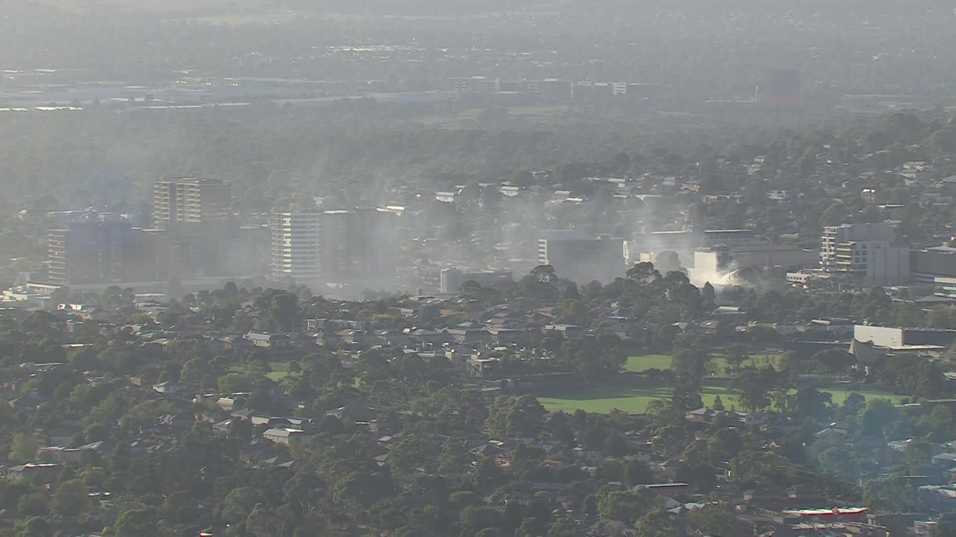 Una vista aérea del humo blanco que se eleva sobre edificios altos, árboles y óvalos durante el día.