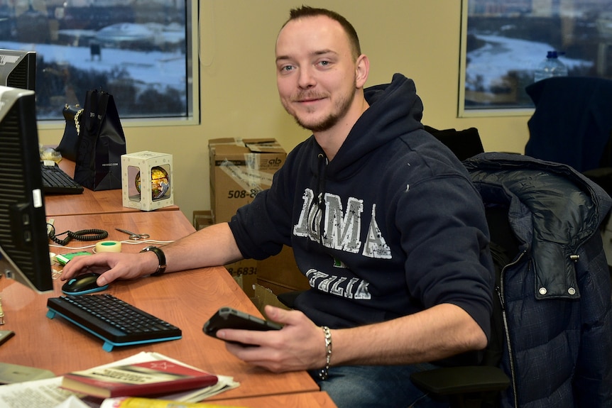 A man holding a phone sits at a desk with a computer and looks at camera.