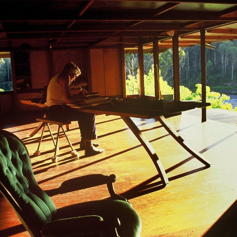 Sun streams in through the structure of a wooden porch at dusk, onto a large wooden desk where Richard Lesplastrier is working