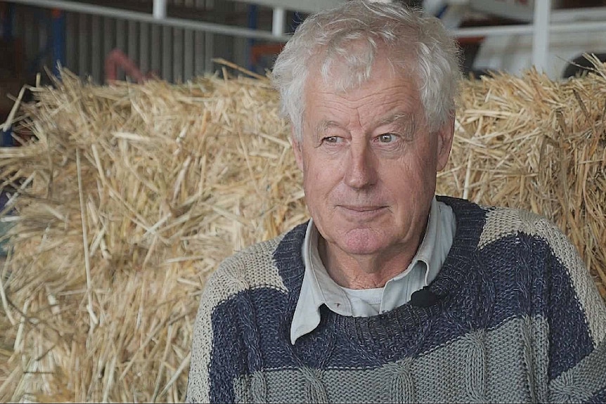 An elderly man poses for a photo inside a hay shed.