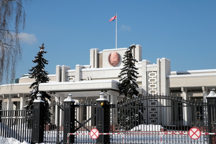 A white building with a flag and embalm on the top and black fence out front.