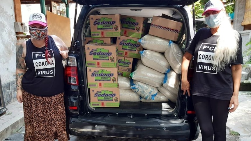 Two Australian women wearing a mask next to a car filled with boxes of instant noodles and rice.