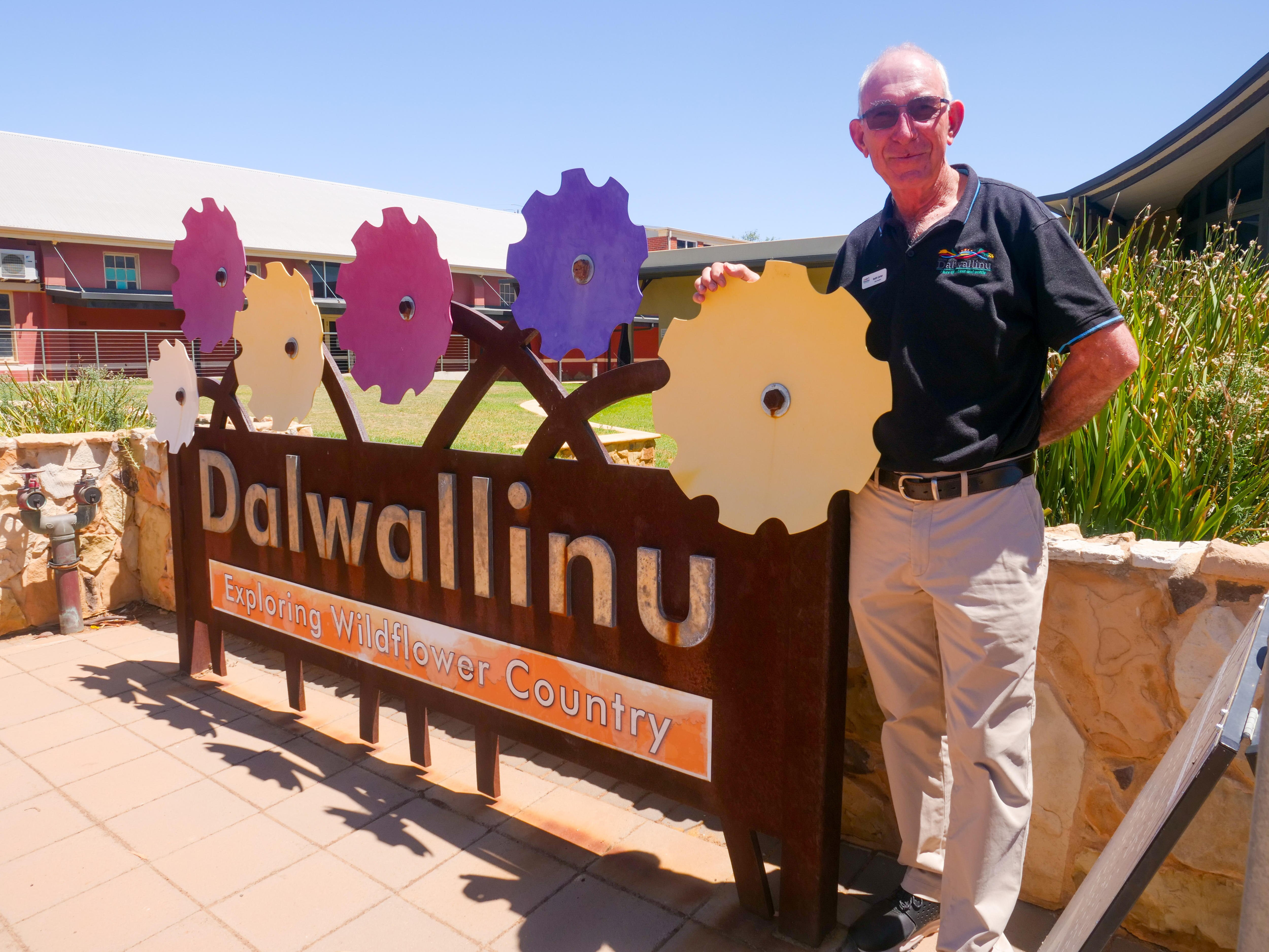 A man standing beside a sign saying 'Dalwallinu' with cut out still flowers above it..