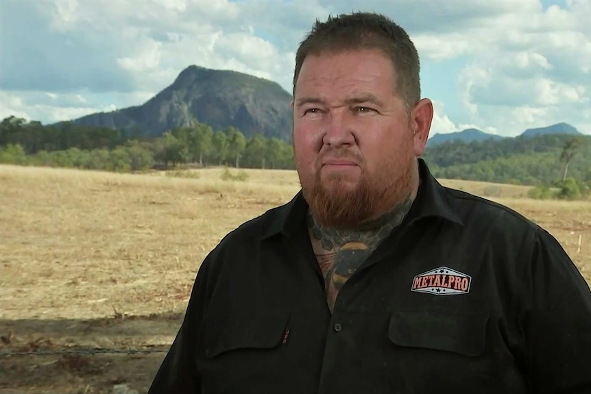 A man wearing a black shirt stands in a field with trees and a mountain in the background.