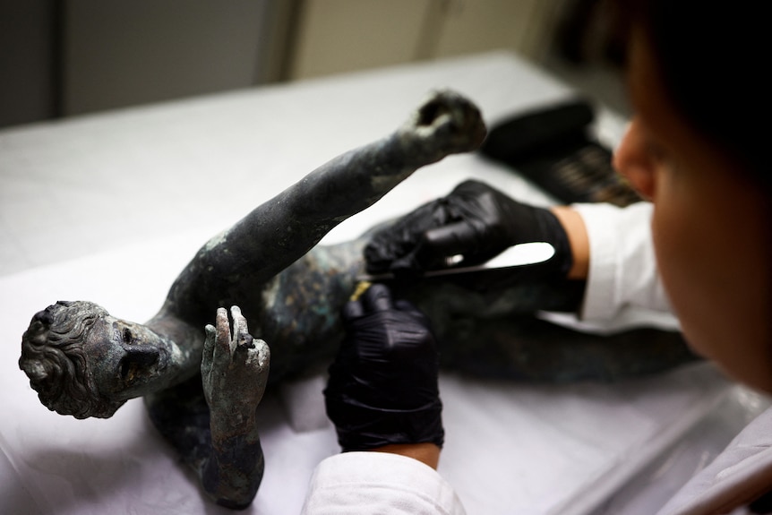 A dark-coloured ancient bronze statue lies on its side on a lab table as a restorer wearing dark gloves works on it.