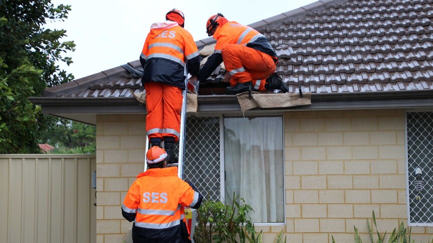 Three SES workers in fluoro gear working to repair a house roof, two of them ion a ladder.