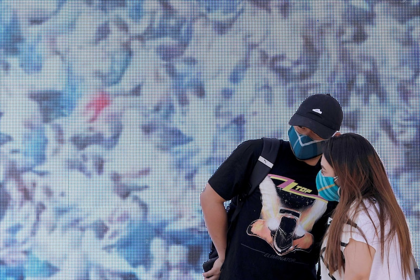 Two young people wearing face masks look off to the side in front of a colourfully painted wall.