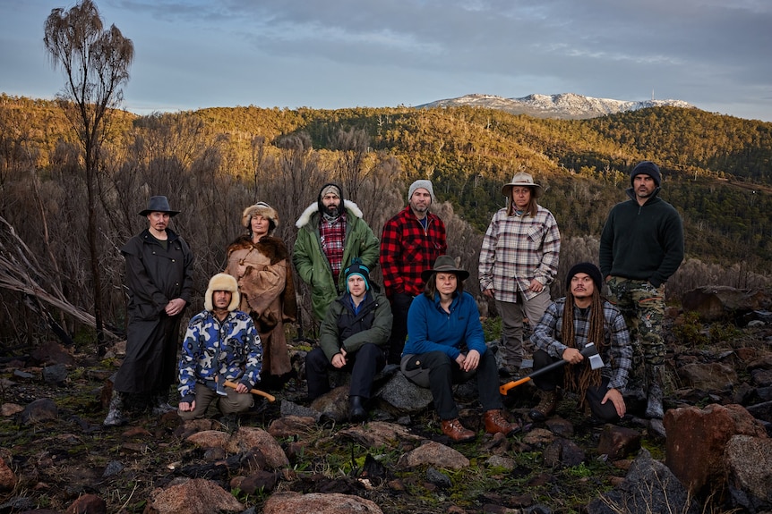 6 people standing and 4 kneeling, in winter weather outdoor gear, rocks in the foreground and forested hills in the background.