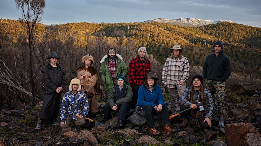 6 people standing and 4 kneeling, in winter weather outdoor gear, rocks in the foreground and forested hills in the background.