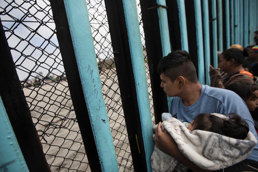 A member of the Central American migrant caravan, holding a child, looks through the border wall to people on the other side.