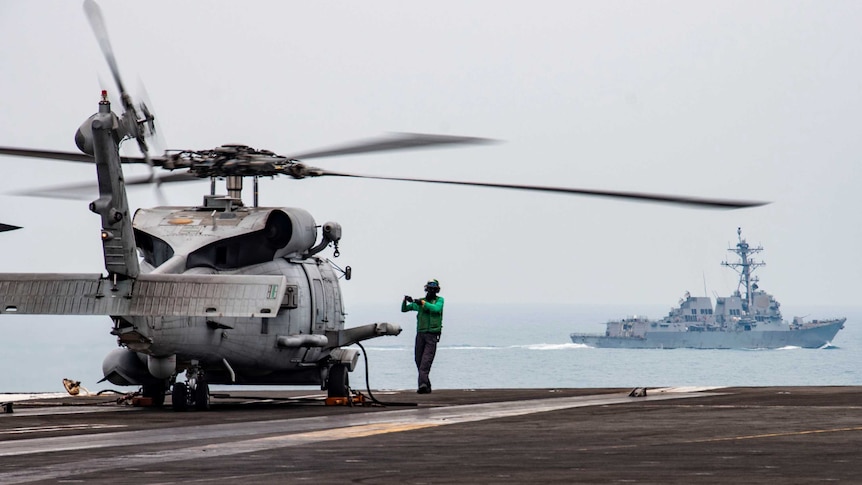 A man in green stands in front of a helicopter on an aircraft carrier with a naval ship in distance.