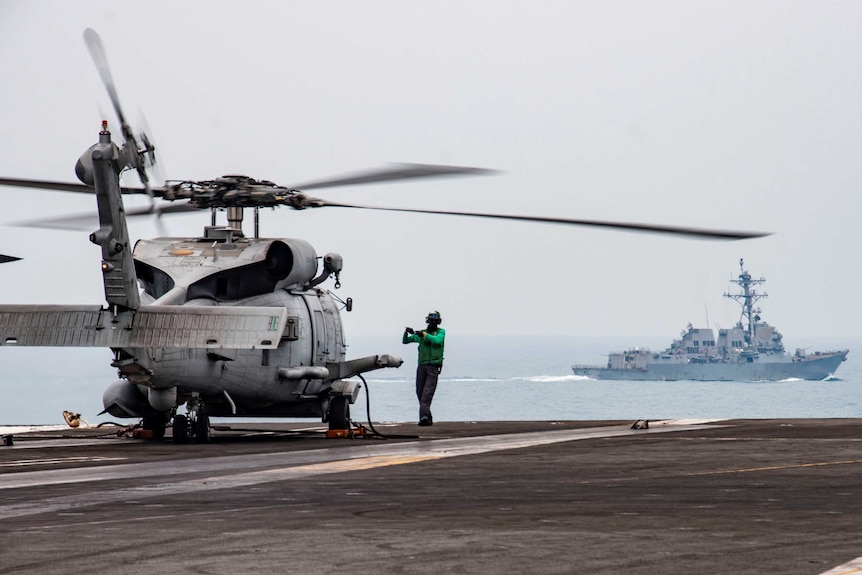 A man in green stands in front of a helicopter on an aircraft carrier with a naval ship in distance.